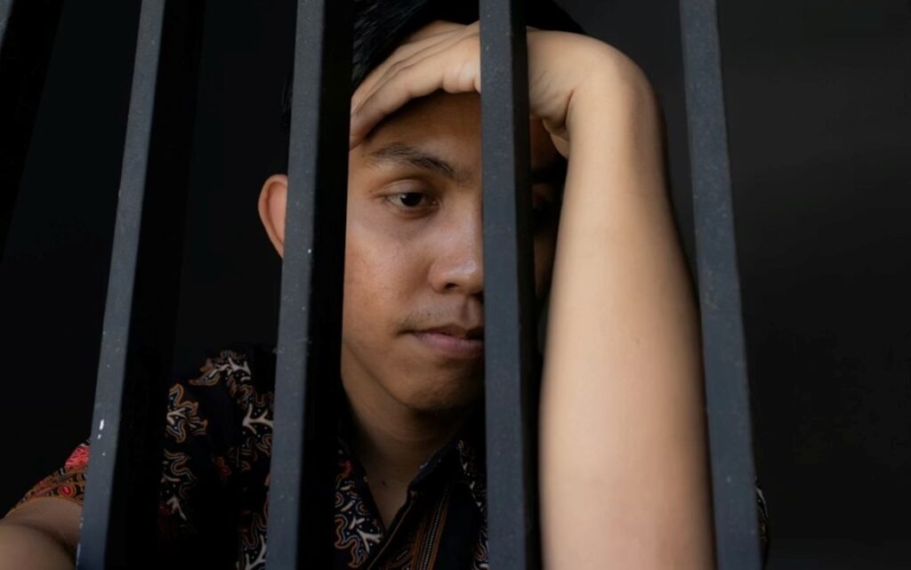 Close-up expression of a man holding bars in prison, looking sad